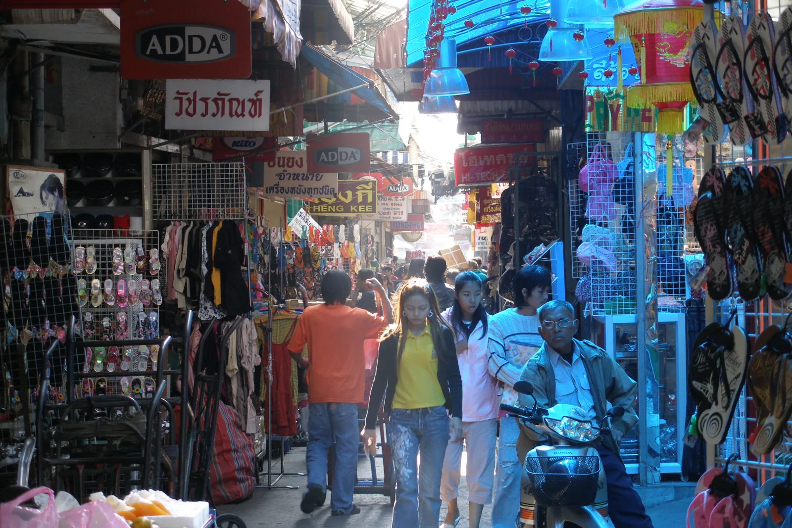 a busy lane in Bangkok with local shops, signs, and people moving through the street